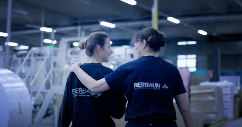 Zwei Mitarbeiterinnen in Arbeitskleidung gehen Arm in Arm durch die Produktionshalle